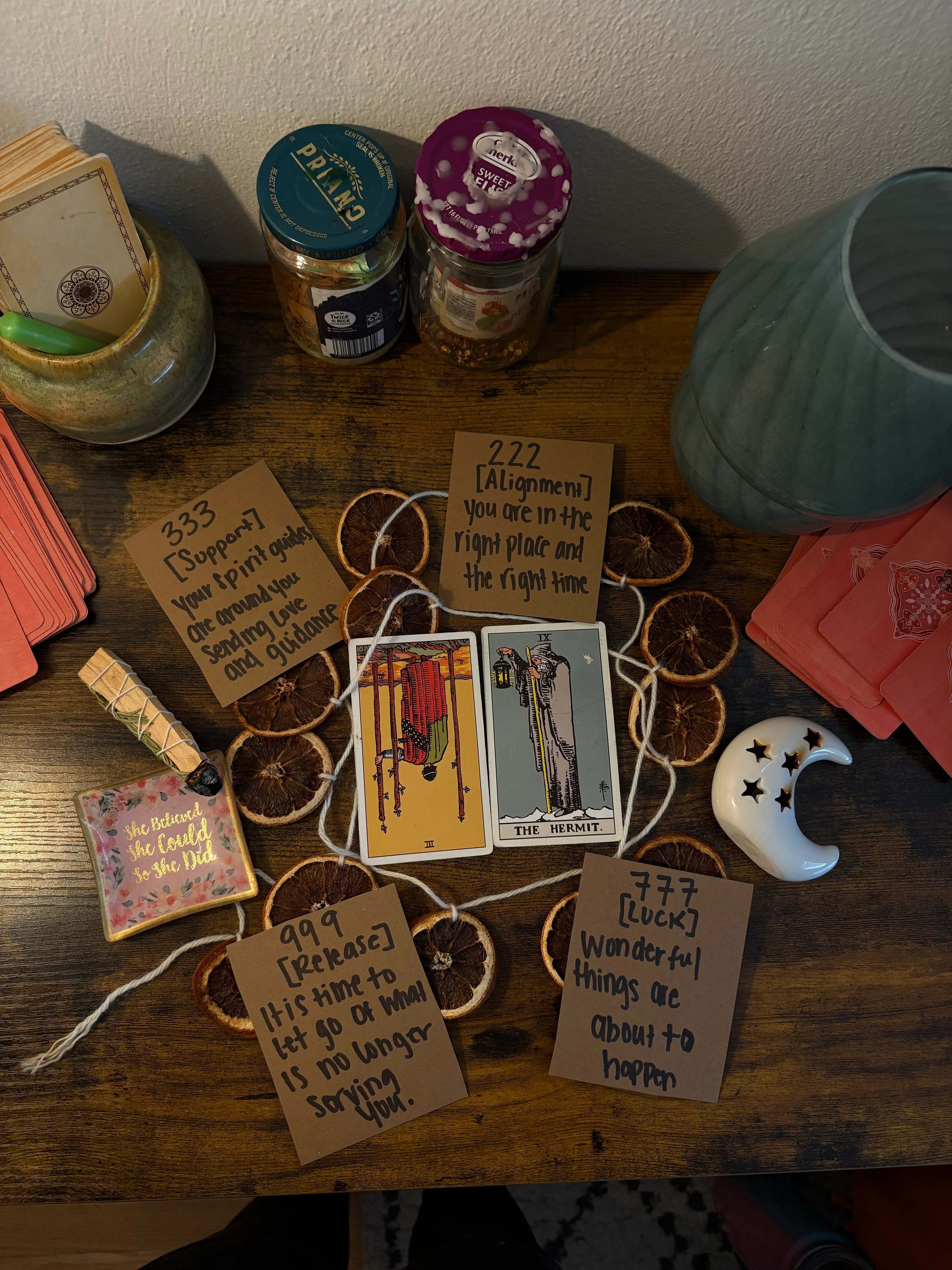 Two tarot cards on a dark wooden desk, surrounded by dried orange, pink cards, palo santo and a ceramic moon.