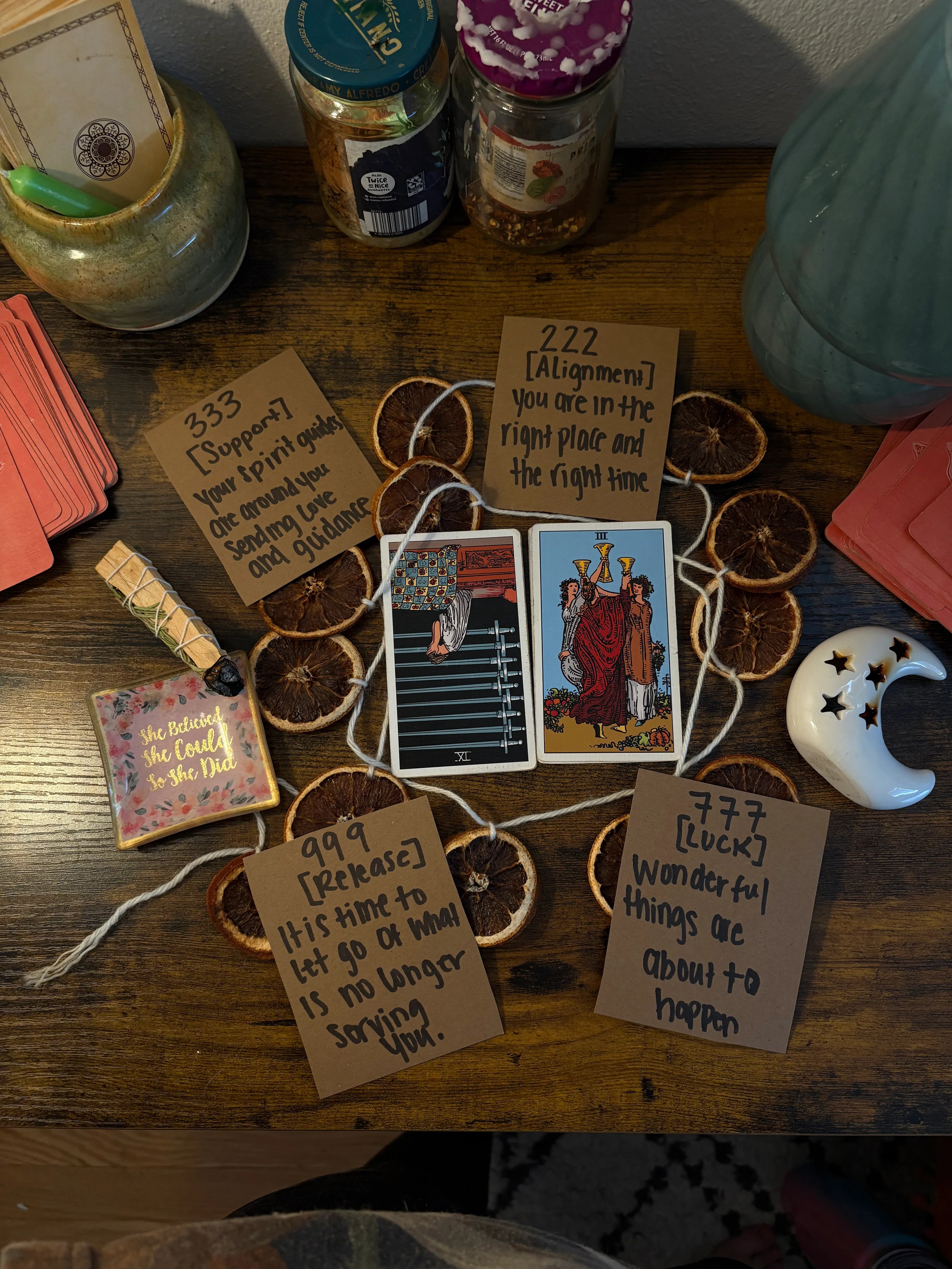 Two tarot cards on a dark wooden desk, surrounded by dried oranges, pink cards, palo santos and a ceramic moon.