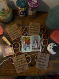 Two tarot cards on a dark wooden desk, surrounded by dried oranges, pink cards, palo santos and a ceramic moon.