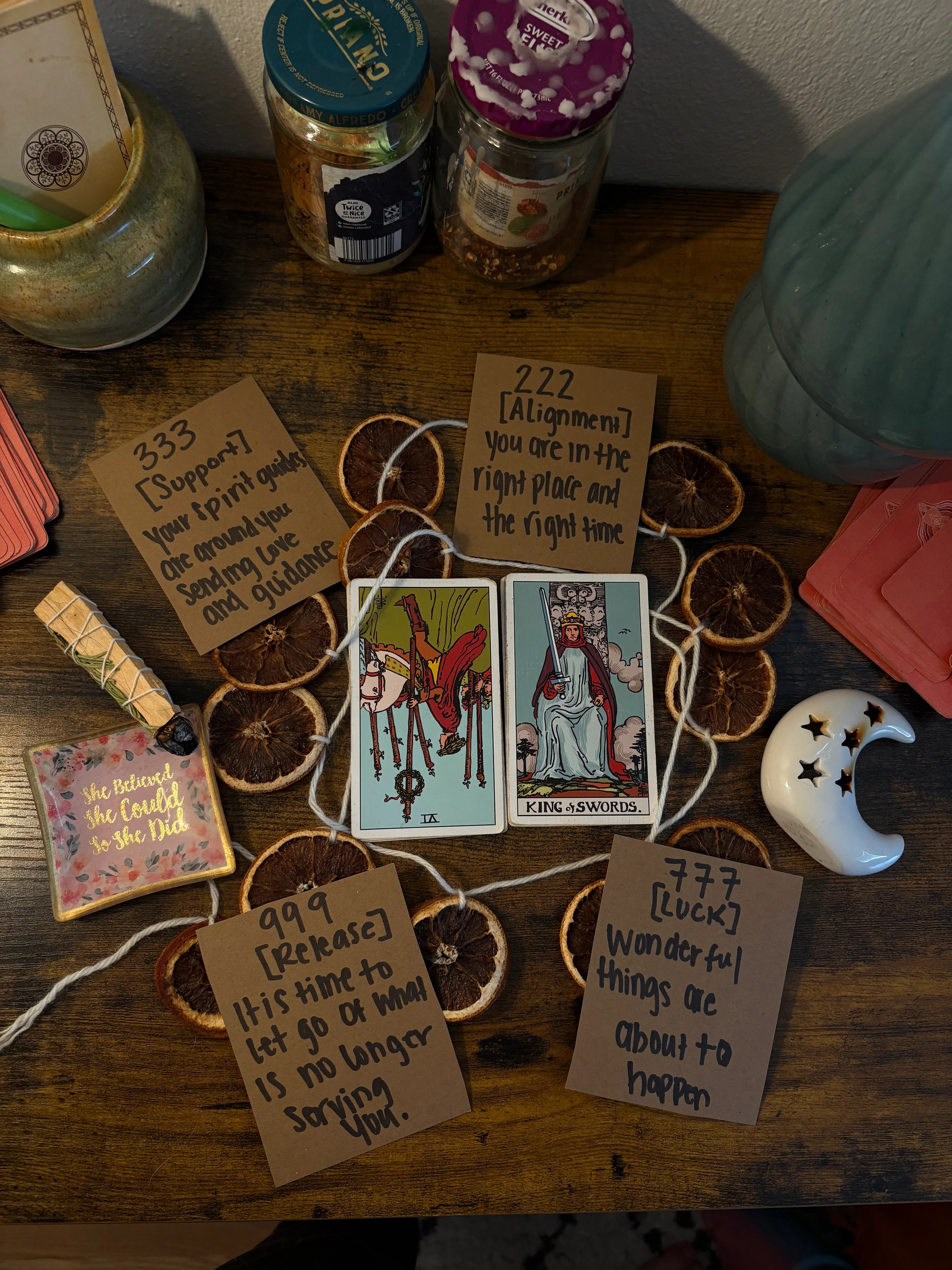 Two tarot cards on a dark wooden desk, surrounded by dried oranges, pink cards, palo santos and a ceramic moon.
